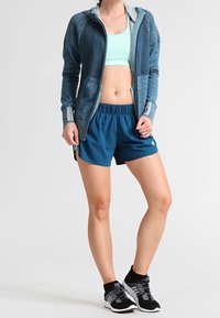 Sininen urheiluhuppari, jossa on harmaa sisäosa, yhdistettynä teal-värisiin shortseihin ja mustiin lenkkareihin. Asussa on tyylikäs muotoilu ja toiminnalliset yksityiskohdat.