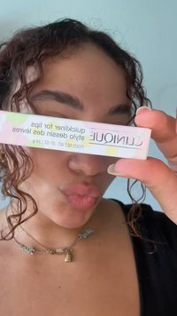Femme aux cheveux bouclés tenant le produit Clinique quickliner pour les lèvres près de son visage, faisant la moue, portant un haut noir et des colliers superposés.