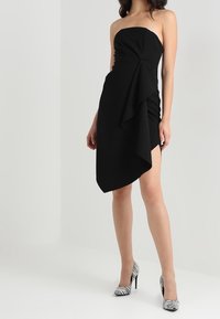Černé mini šaty bez ramínek s drapérií v přední části a vysokým rozparkem, doplněné černobílými pruhovanými jehlovými podpatky.
