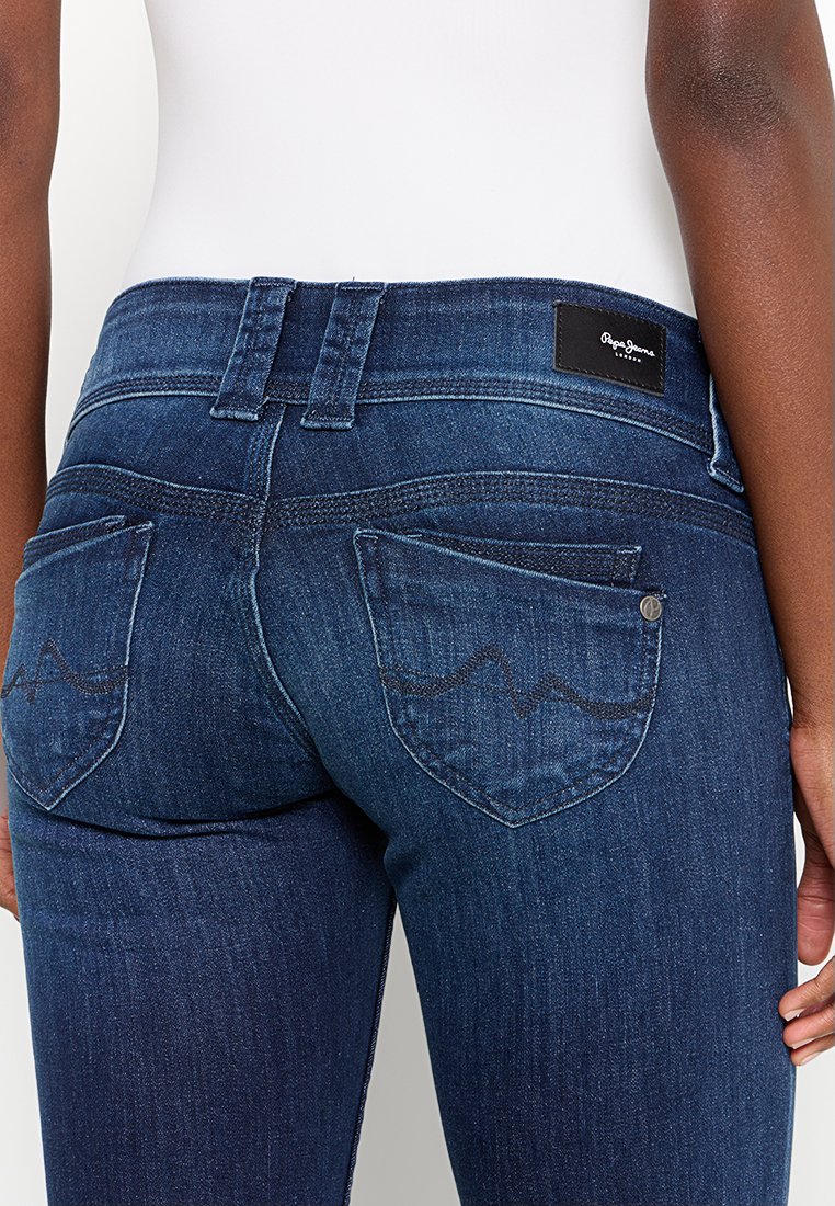 Jeans de mezclilla azul oscuro con un diseño ajustado. Las características incluyen dos bolsillos traseros con costuras en patrón de ola y una etiqueta de cuero de marca.