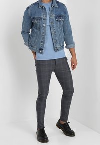 Džínová bunda s klasickým střihem, světle modrá; navržena přes žebrovaný svetřík světle modré barvy. Slim-fit šedé kostkované kalhoty. Černé boty dokončují celý vzhled.