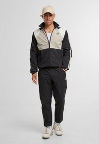 Giacca nera e beige con zip e collo alto, abbinata a pantaloni jogger neri e sneaker bianche. Presenta dettagli con logo sulla giacca.