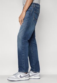 Jeans in denim blu scuro con vestibilità dritta, design a cinque tasche e cuciture a contrasto. Abbinati a sneakers grigio chiaro e viola.