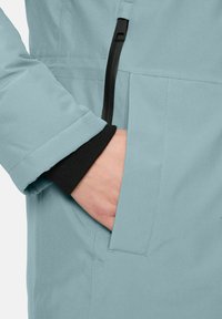 Hand in der Vordertasche einer hellblauen Jacke mit sichtbarem schwarzen Innenbündchen und vertikalem Reißverschluss-Detail.