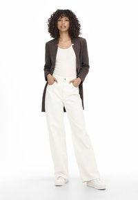 Blazer brown lungo sopra una canotta bianca e jeans bianchi a gamba larga, con una texture liscia e una vestibilità rilassata. Le sneakers completano l'outfit.