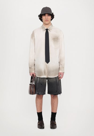 Chemise boutonnée gris clair avec des taches sombres, cravate noire, shorts en denim noir déchirés, sac marron, chaussettes noires, et chaussures marron.