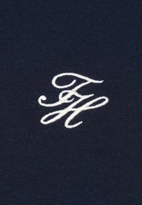 Tengerészkék szövet bordázott textúrával, fehér hímzett monogram logóval, stilizált betűtípussal.