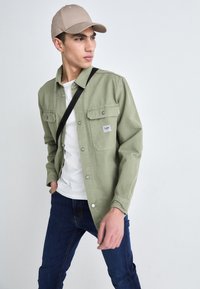 Chemise boutonnée vert clair avec deux poches poitrine, portée par-dessus un t-shirt blanc et assortie à un jean bleu foncé ; comprend une casquette beige.