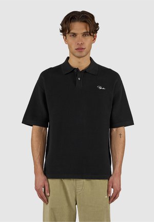 Młody mężczyzna o krótkich falowanych włosach, ubrany w czarną koszulkę polo z krótkim rękawem i beżowe spodnie, stoi na jednolitym szarym tle.