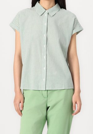 Kvinde iført en kortærmet, grøn og hvid lodret striped skjorte med krave og knapper, kombineret med lysegrønne bukser.