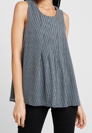 Femme portant un haut sans manches, ample avec un petit motif géométrique noir et blanc et un pantalon noir sur fond uni.