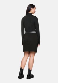Schwarzes Kleid mit langen Ärmeln, figurbetontem Schnitt und weißem Akzentgürtel. Strukturierter Stoff und knielanges Design, kombiniert mit Ankle Boots.