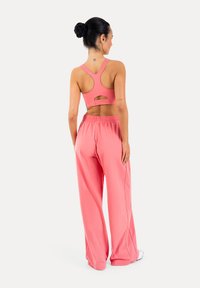 Donna in piedi di spalle che indossa un reggiseno sportivo rosa e pantaloni rosa larghi, capelli legati in uno chignon, su sfondo bianco.