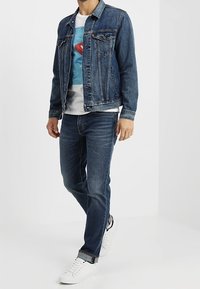Jeansjacka med två framfickor, medelhavsblå färg; bärs över en vit T-shirt med grafisk design; matchas med mörka jeans och vita sneakers.