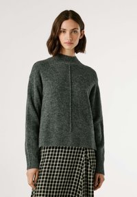 Suéter de malha canelada cinza escuro com gola alta, apresentando um detalhe de costura central e mangas longas, combinado com uma saia xadrez preto e branco.