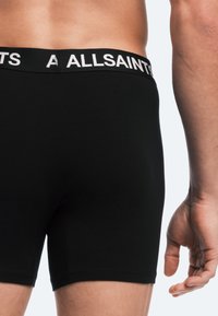 Schwarze Boxer-Brief mit weichem Material, versehen mit einem weißen "ALLSAINTS"-Logo am elastischen Bund. Nahaufnahme des Rückens und des Hüftbereichs.