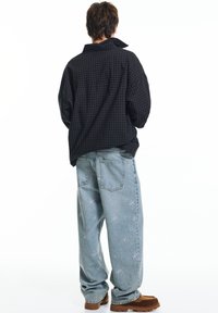 Zwarte geruite oversized blouse, lichtblauwe spijkerbroek met bloemenborduursel en bruine suède schoenen. Model met de rug naar de camera.