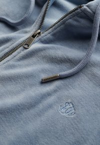 Sudadera con cremallera de color azul claro, fabricada en un tejido suave, con una superficie texturizada, logo bordado y cremallera de metal en contraste.