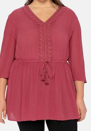 Femme portant un chemisier rose poudré à manches trois-quarts, avec un col en V orné de dentelle et une ceinture à nouer avec des pampilles.