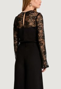Kobieta ubrana w czarny strój z kwiatową koronką na długich rękawach i plecach, zestawiony z czarnymi spodnie o wysokim stanie, stojąca bokiem.