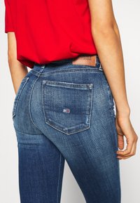Persona que lleva puestos jeans de mezclilla mid-blue de Tommy Jeans con bolsillos traseros y una camisa roja metida por dentro, mostrado desde la cintura hacia abajo.