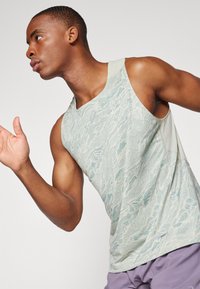 Débardeur de sport sans manches vert clair avec un motif de vagues texturé et des accents en mesh aux épaules, associé à un short violet.