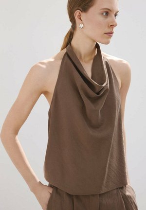 Femme portant un top marron sans manches drapé à col halter et un pantalon assorti, avec des boucles d'oreilles clip argentées et les cheveux en queue de cheval basse.