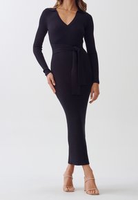 Kvinna klädd i en lång svart ribbad stickad klänning med V-ringning och knytväska, kombinerad med nudefärgade högklackade sandaler, stående mot en enfärgad bakgrund.