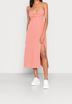 Femme portant une robe midi rose sans manches avec une fente devant et des baskets blanches compensées, debout devant un fond uni.