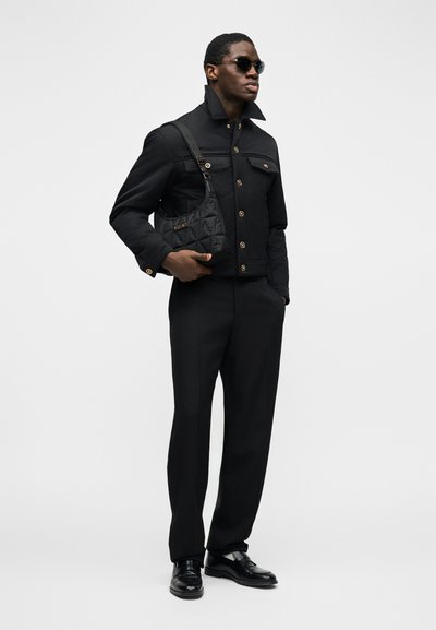 Άνδρας που φοράει μαύρο σακάκι με κουμπιά, μαύρο παντελόνι, μαύρα μοκασίνια, γυαλιά ηλίου, κρατώντας μια μικρή μαύρη καπιτονέ τσάντα ώμου.