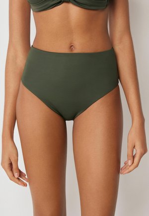 Frau trägt olivgrüne Bikinihose mit hohem Bund und passendem Oberteil, steht vor einem schlichten hellen Hintergrund.