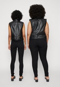 Colete de pele preto com design sem mangas, fechos de botão e textura suave, usados sobre leggings pretas. Ambos os modelos estão de costas para a câmara.