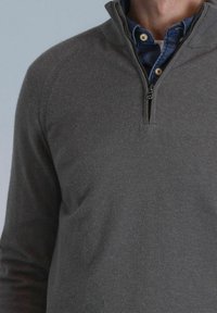 Grijze pullovertrui met een half-rits kraag, gemaakt van zacht materiaal, met geribbelde schoudernaden en gelaagd over een blauwe jeanshemd.