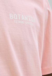 Jasnoróżowa bawełniana koszulka z krótkim rękawem, z napisem "BOTANICA CITRUS SPECIAL" w białym druku na przodzie.