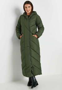 Długa, zielona kurtka puchowa z kapturem, z zamkiem z przodu, diagonalnym wzorem pikowania i bocznymi kieszeniami. Noszona z czarnymi botkami sięgającymi kostki.