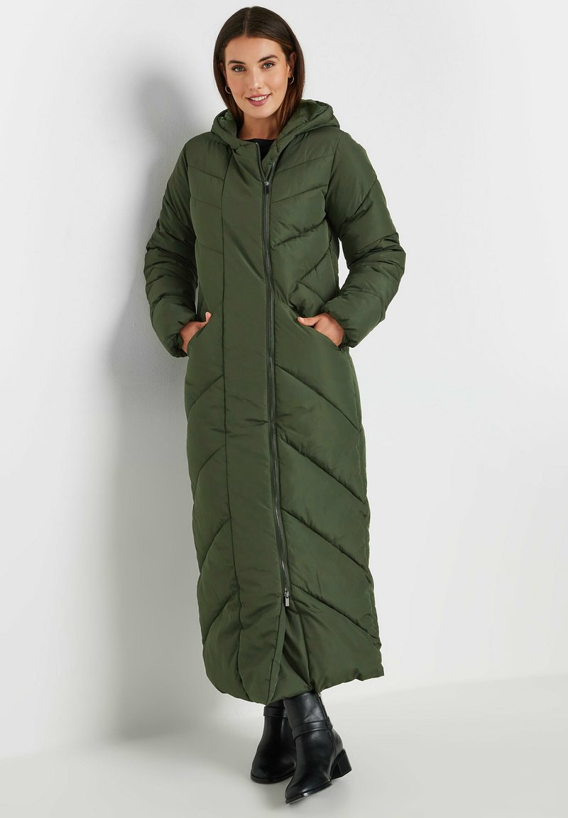 Długa, zielona kurtka puchowa z kapturem, z zamkiem z przodu, diagonalnym wzorem pikowania i bocznymi kieszeniami. Noszona z czarnymi botkami sięgającymi kostki.