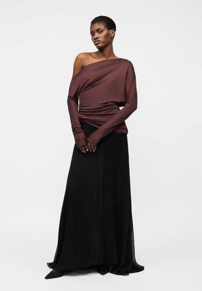 Kobieta stoi, ubrana w brązowy top z odkrytymi ramionami i długim rękawem oraz czarną, długą, zwiewną spódnicę do ziemi na tle jednolitego białego tła.