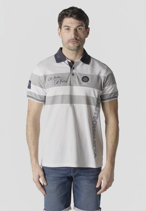 Hombre con camiseta polo a rayas blancas y grises con cuello azul marino y pantalones cortos de mezclilla, de pie contra un fondo claro y liso.