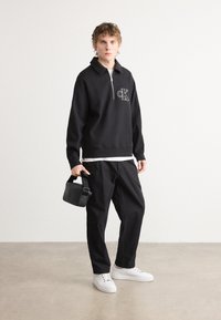 Black zip-up pullover with logo, paired with black pants and white sneakers. Model holds a small black bag. Smooth texture and casual fit.