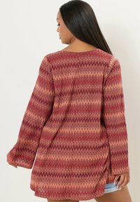 Cardigan a maniche lunghe con motivo a zigzag in rosso, rosa e beige. Tessuto leggero, vestibilità comoda e spacchi laterali per facilitare i movimenti.