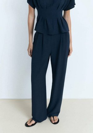 Femme portant un pantalon bleu marine ample taille haute et un haut ajusté à manches courtes assorti, debout avec des sandales noires sur un sol clair.