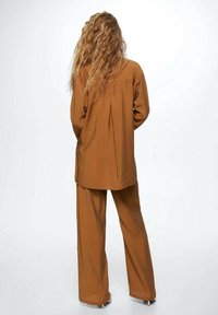 Bruin tweedelig outfit bestaande uit een losvallend lang shirt en wijde broek, gemaakt van gestructureerde stof, met een rugplooidetail.