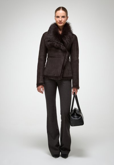 Chaqueta de ante marrón con cuello de piel, diseño ajustado y detalles con cremallera. Combinada con pantalones de campana grises y un bolso negro.