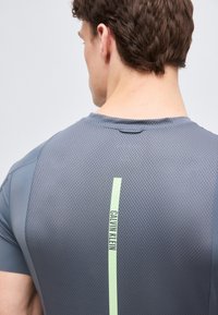 Άνδρας με γκρι υφασμάτινη αθλητική μπλούζα με κάθετη ανοιχτοπράσινη ρίγα και το κείμενο "Calvin Klein" στην πάνω πλάτη.