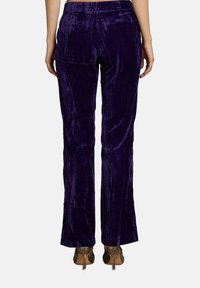 Pantaloni in velour viola con una forma leggermente svasata, caratterizzati da una finitura testurizzata e una cintura liscia, indossati con tacchi aperti.