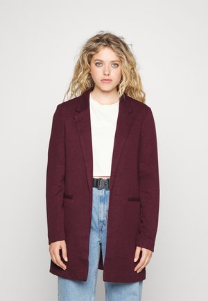 Burgundy long blazer with a sharp lapel, two front pockets, and a relaxed fit, worn over a cream T-shirt and high-waisted jeans.