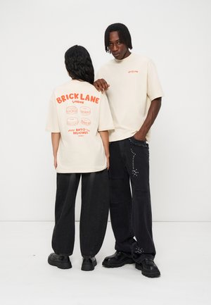 To modeller har cremefarvede t-shirts med orange "Brick Lane London" tekst; den ene viser forsiden, den anden bagsiden med madillustrationer.