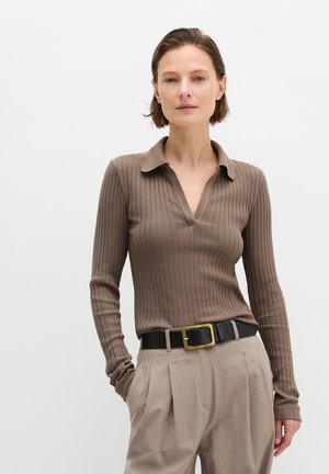 Kobieta z krótkimi włosami, ubrana w brązową prążkowaną bluzkę z długim rękawem, beżowe plisowane spodnie i czarny pasek ze złotą klamrą, z rękami w kieszeniach.