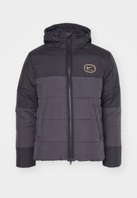 Svart dunjacka med huva, kontrasterande grå paneler, framknäppning med dragkedja och en guldfärgad logo på bröstet. Har vaddering med kviltade mönster.