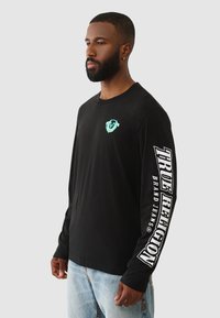 Schwarzes Langarm-T-Shirt mit einem kreisförmigen blauen Logo auf der Brust und dem Text "TRUE RELIGION" in Weiß auf dem linken Ärmel.